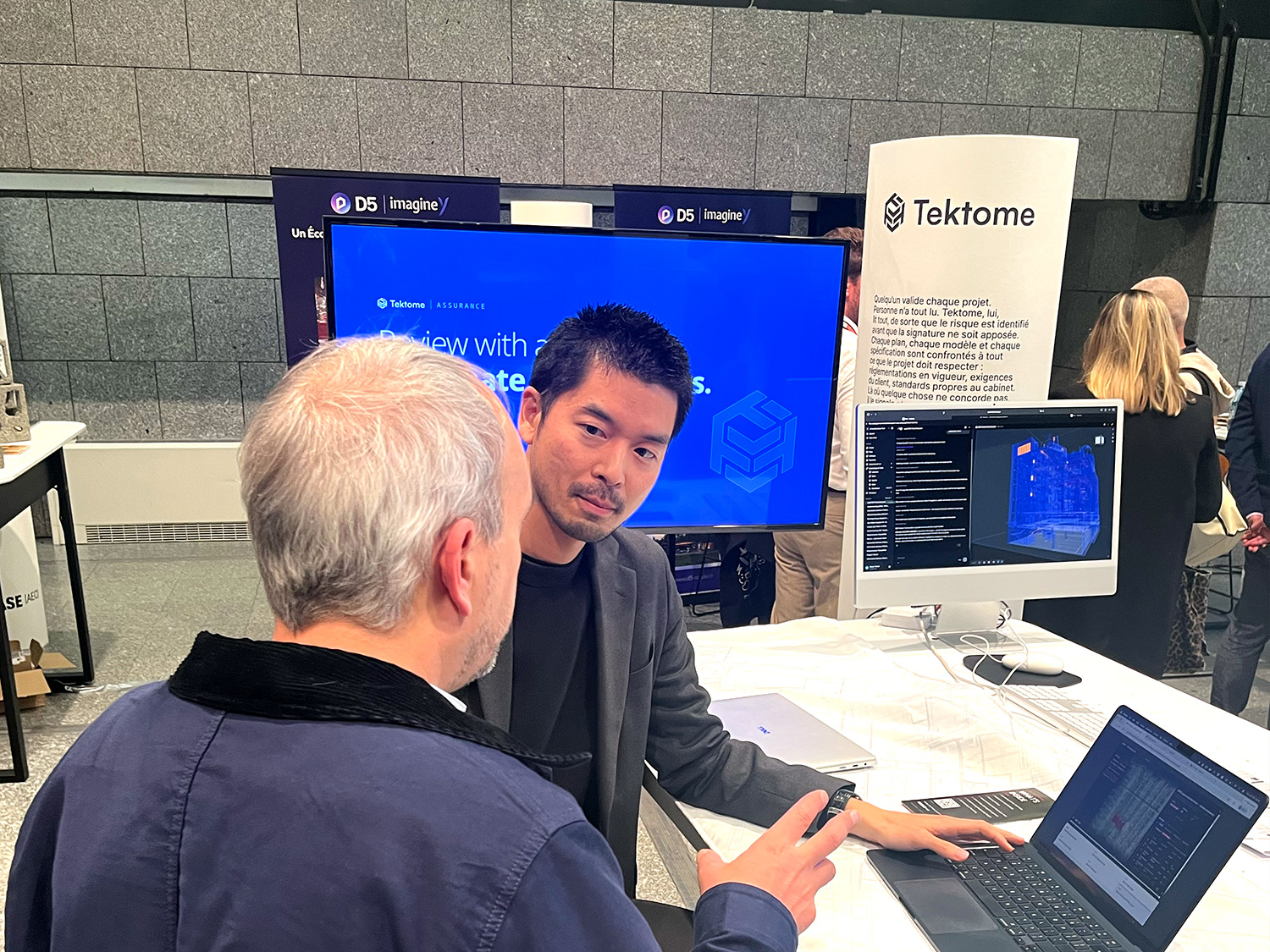 RELEASE [AEC] 2026 のTektomeブース。大型スクリーンとBIMモデルを映したディスプレイの前で、スタッフが来場者とノートPCの画面を見ながら話している